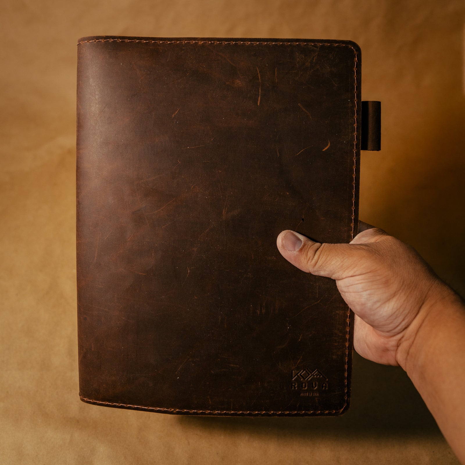 Hand holding a dark brown leather notebook cover against a beige background