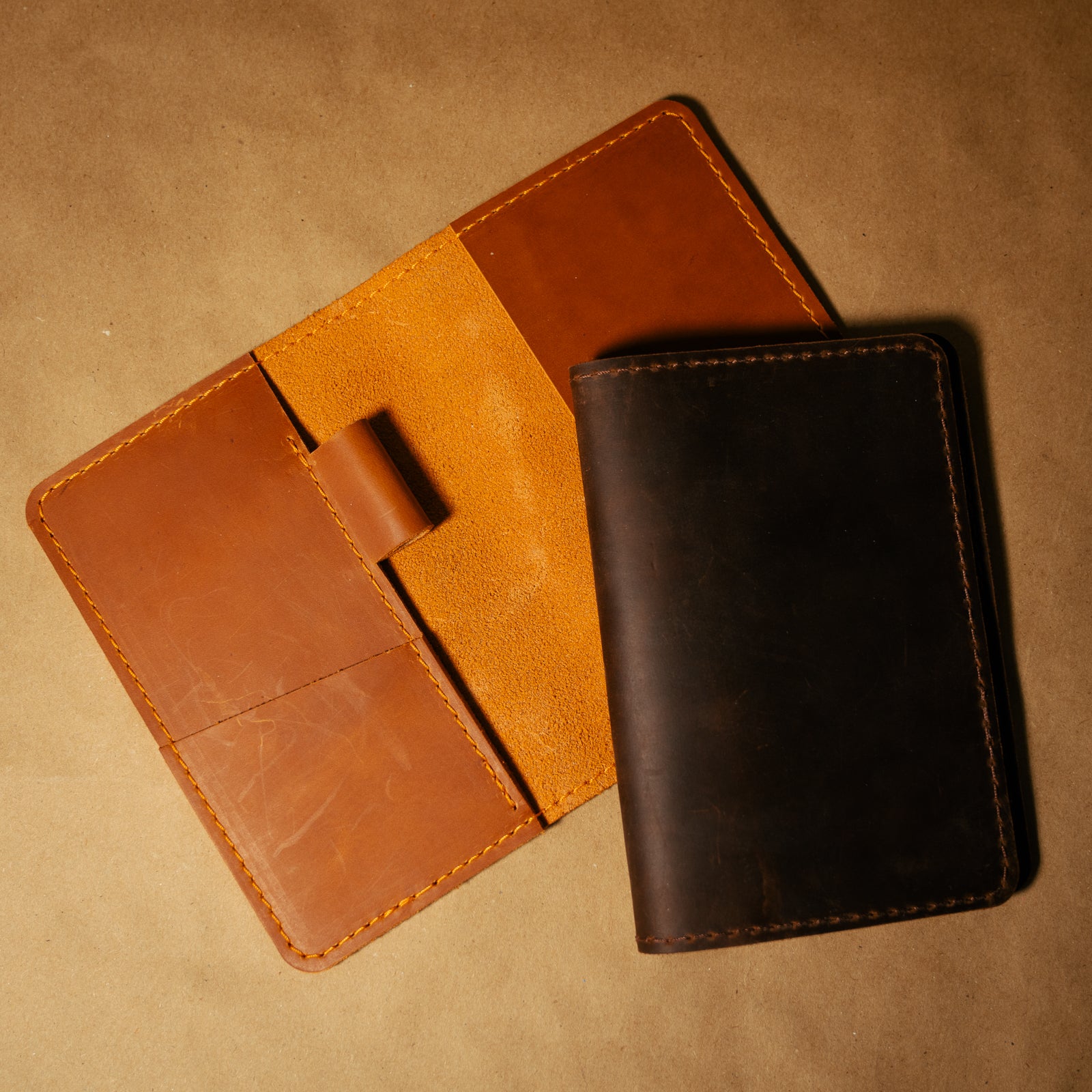 Two leather field notes journal covers, one dark brown and one whiskey orange, on a beige background