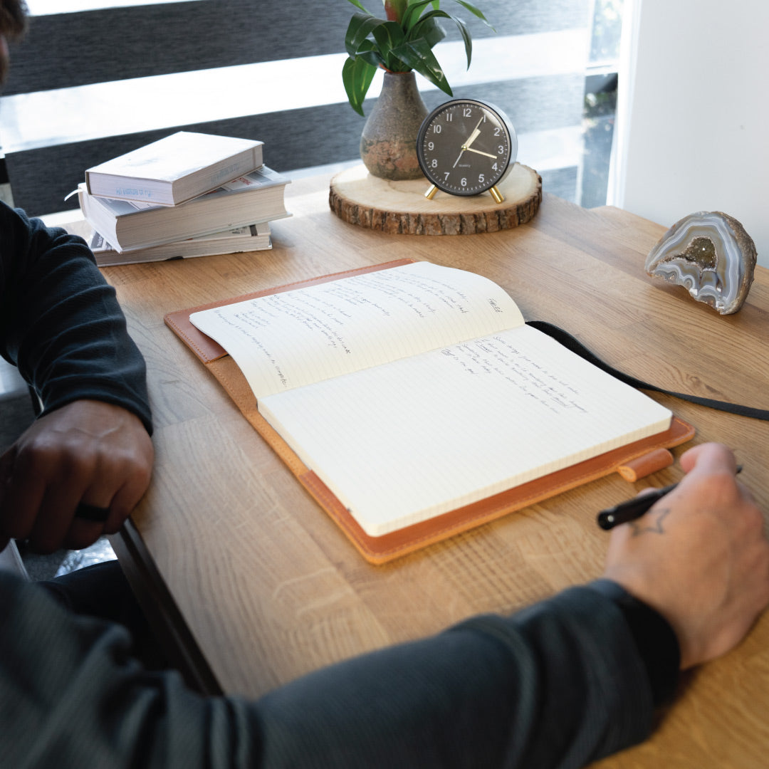 Person writing in a notebook on a wooden desk with books, a clock, and a plant in the background.
