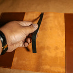 Hand holding a black leather bookmark over a whiskey orange leather journal cover