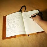 Handwriting in a Moleskine notebook with leather cover and a hand holding a pen on a wooden surface