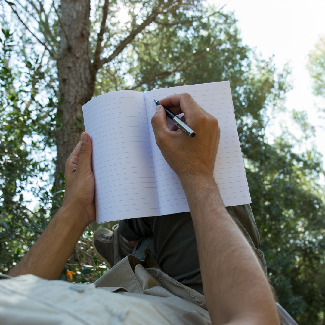 Person writing in a field notes notebook with trees in the background